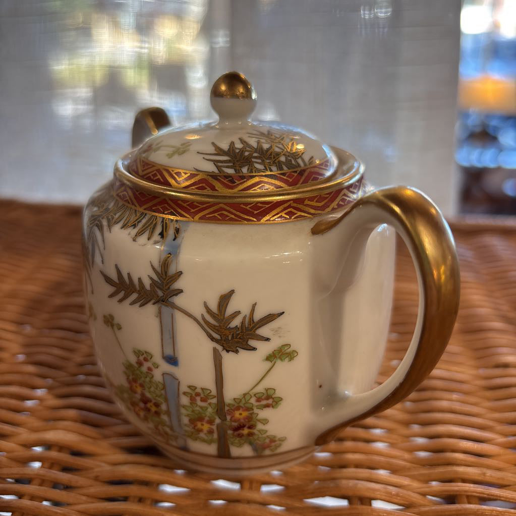 A ceramic lidded sugar bowl with painted floral elements on a rattan self in a room setting. 
