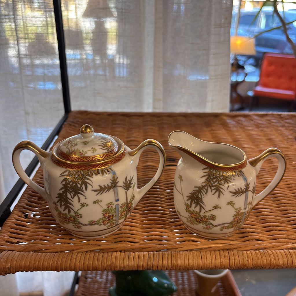 A ceramic lidded sugar bowl and creamer on a rattan self in a room setting. 