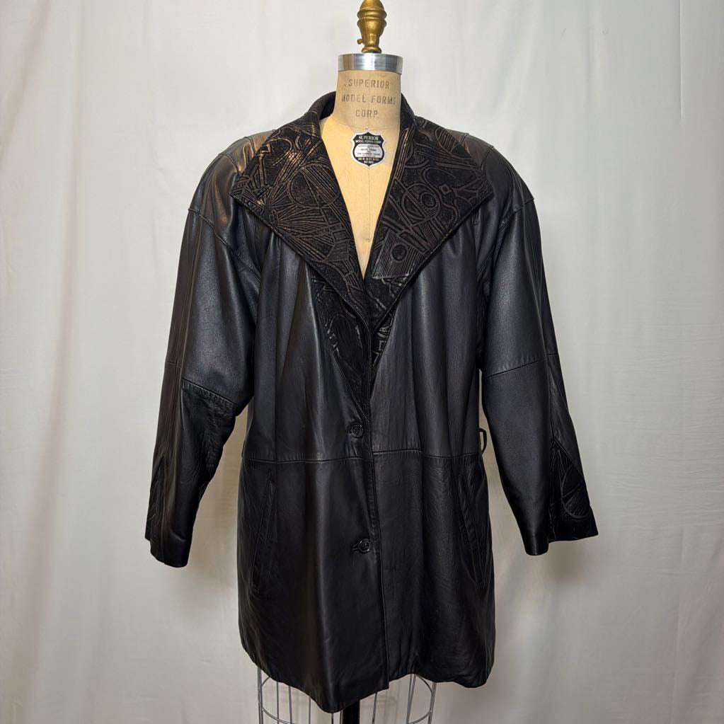 A black leather trench coat with wide shoulders and an abstract design on the lapels. Its on a mannequin in front of a white wall. 