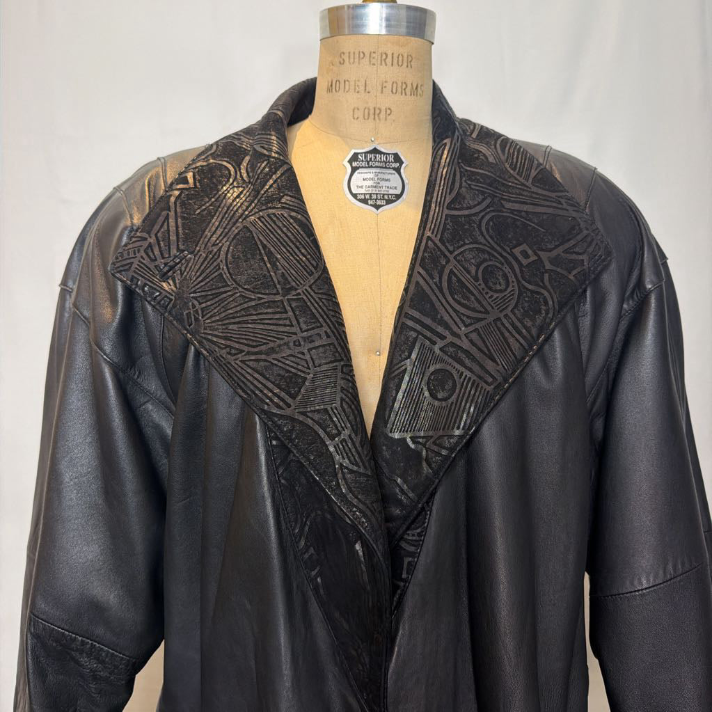 A black leather trench coat with wide shoulders and an abstract design on the lapels. Its on a mannequin in front of a white wall. 