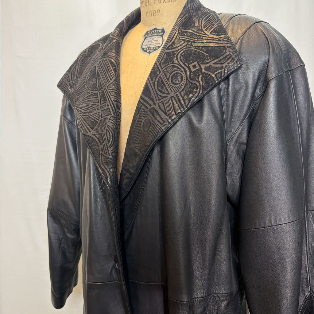 A black leather trench coat with wide shoulders and an abstract design on the lapels. Its on a mannequin in front of a white wall. 
