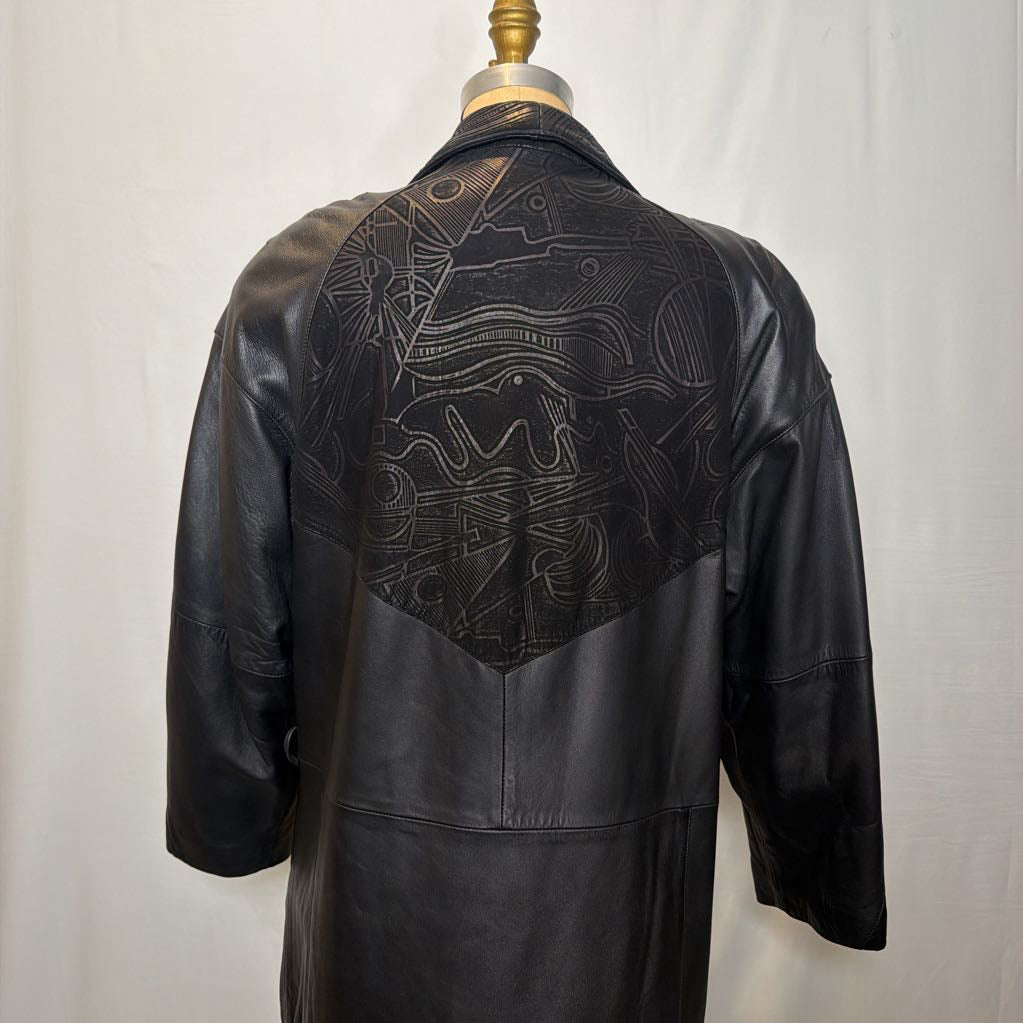 A black leather trench coat with wide shoulders and an abstract design on the back. Its on a mannequin in front of a white wall. 