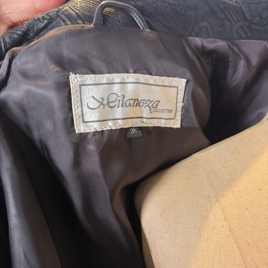A black leather trench coat with wide shoulders and an abstract design on the lapels. Its on a mannequin in front of a white wall. 