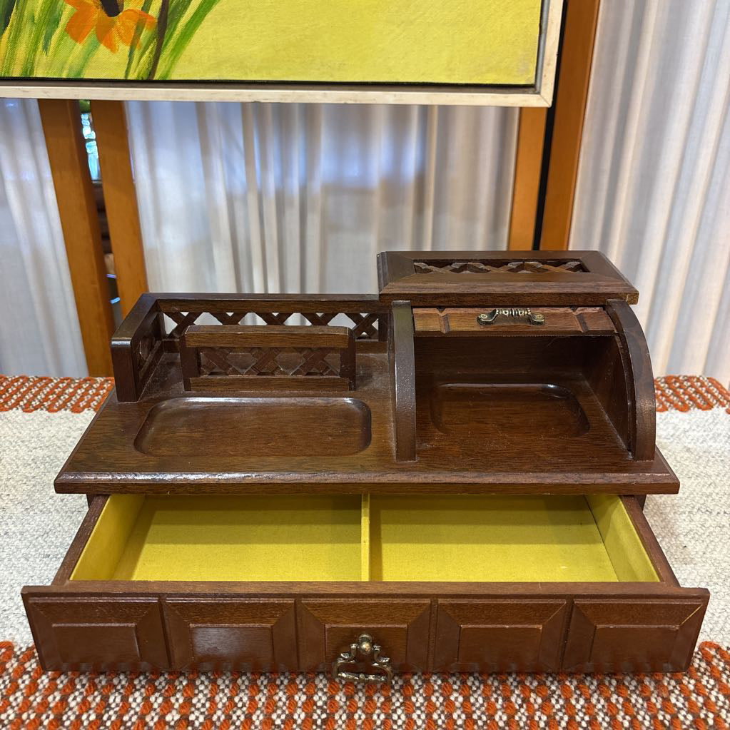 An open wooden jewelry box/valet box on a woven runner in front of a white screen with part of a yellow painting showing.