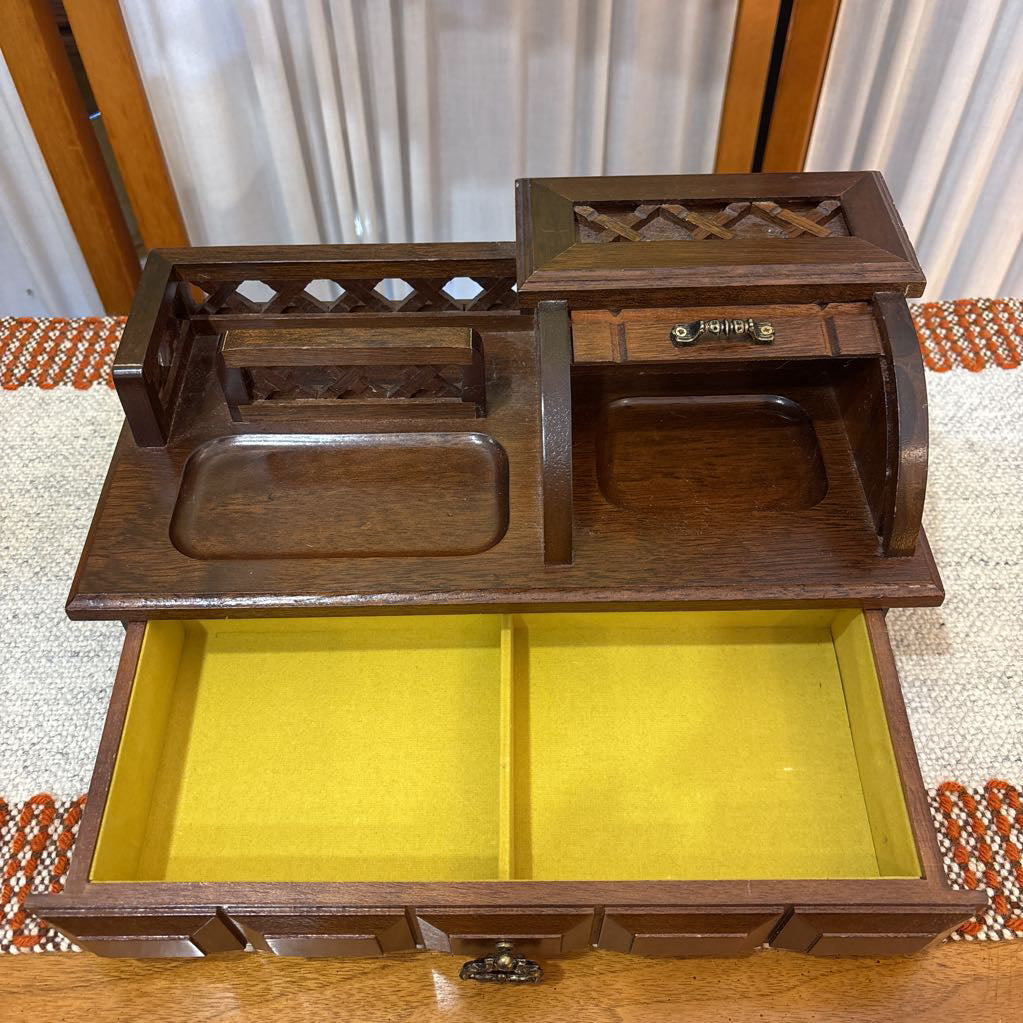 An open wooden jewelry box/valet box on a woven runner in front of a white screen with part of a yellow painting showing.