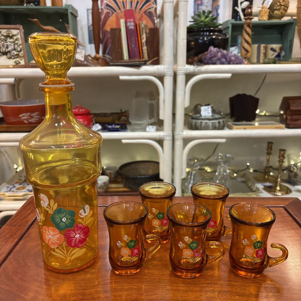 An amber glassware set with a decanter and 5 cordial glasses all with a floral pattern on the front sitting on a wooden table in a store setting. 
