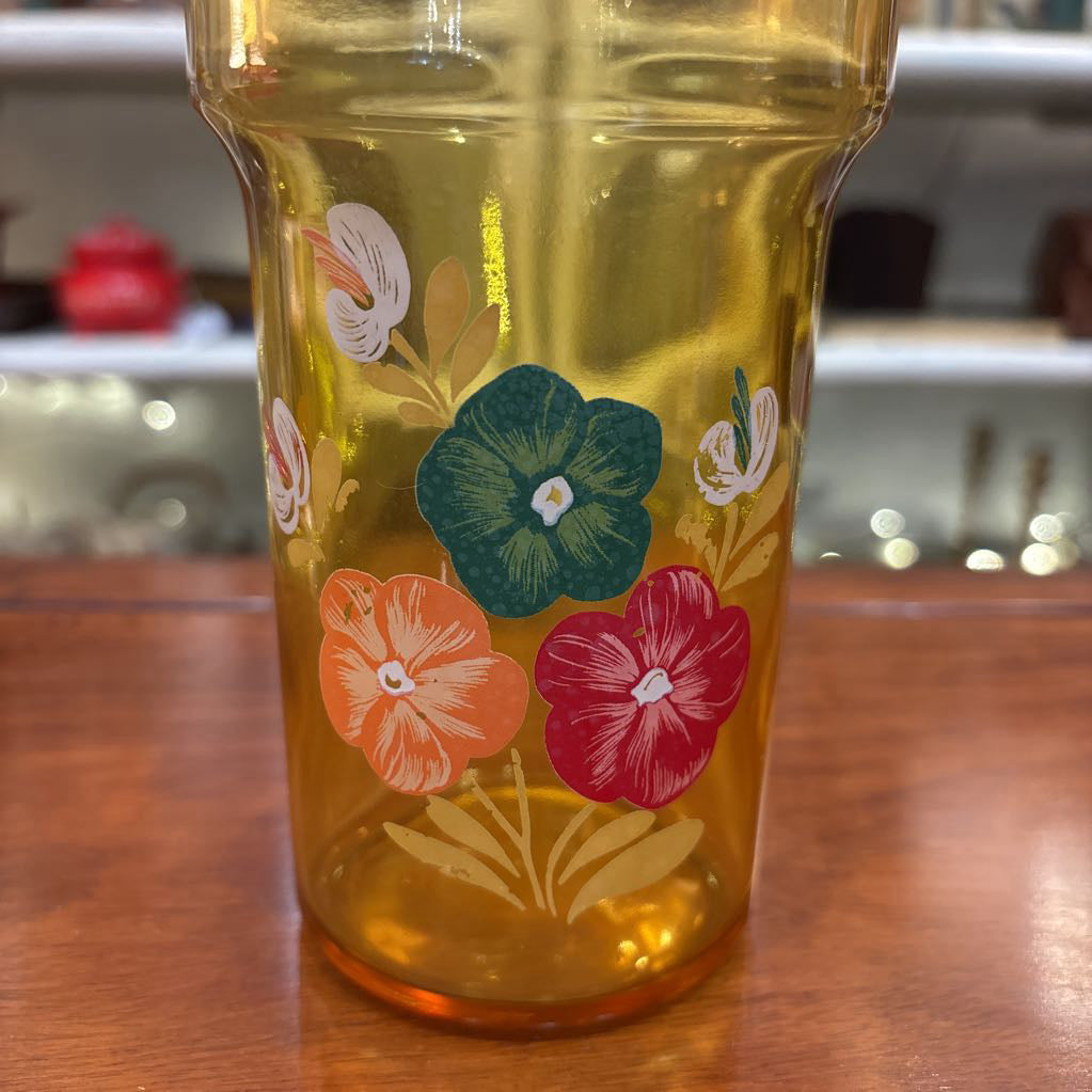 An amber glassware set with a decanter and 5 cordial glasses all with a floral pattern on the front sitting on a wooden table in a store setting. 