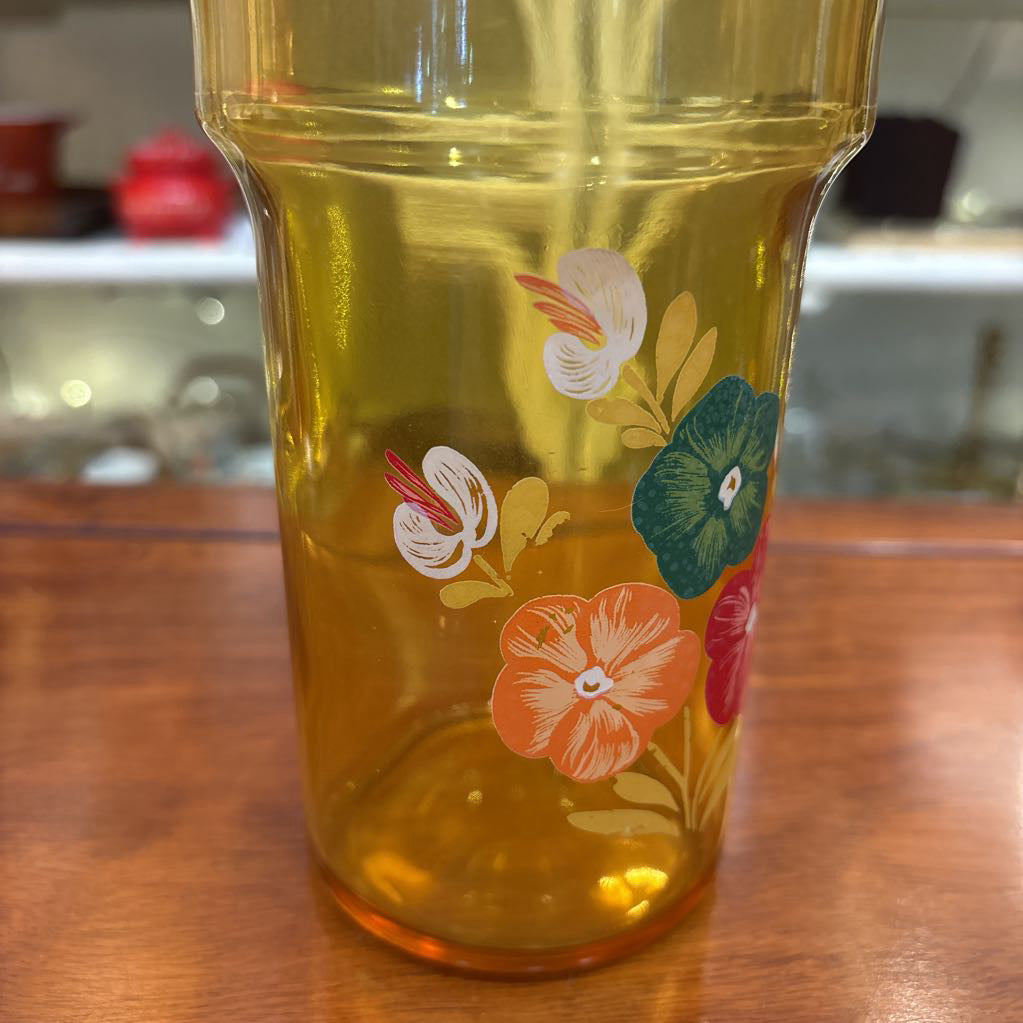 An amber glassware set with a decanter and 5 cordial glasses all with a floral pattern on the front sitting on a wooden table in a store setting. 