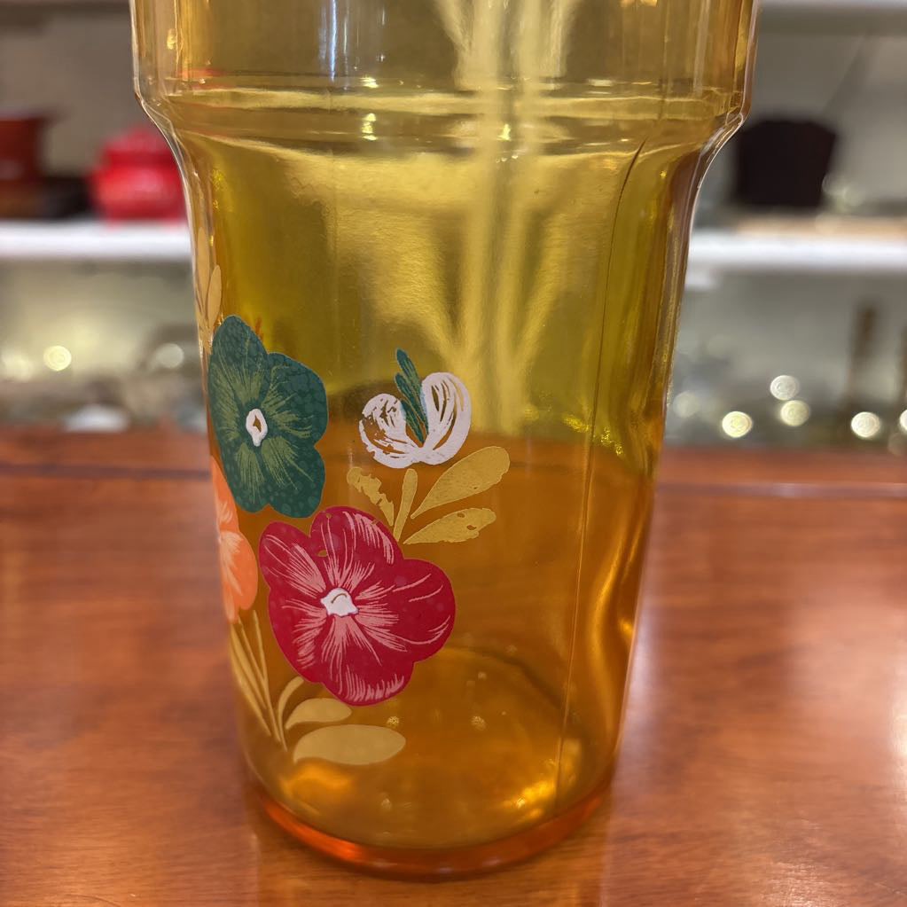 An amber glassware set with a decanter and 5 cordial glasses all with a floral pattern on the front sitting on a wooden table in a store setting. 