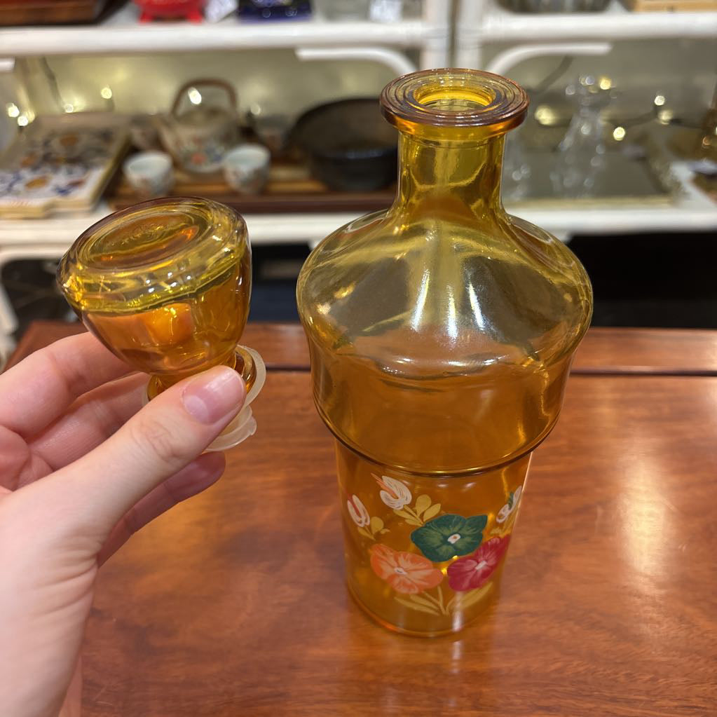 An amber glassware set with a decanter and 5 cordial glasses all with a floral pattern on the front sitting on a wooden table in a store setting. 