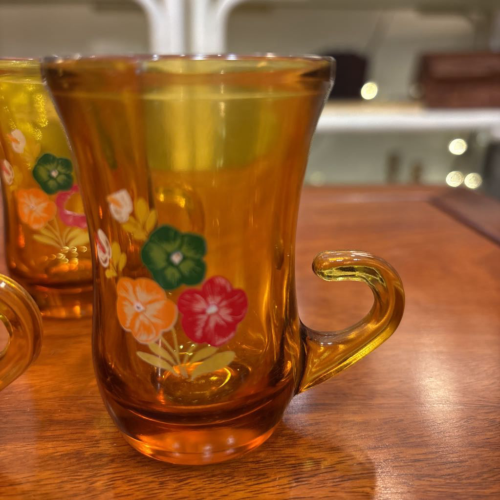 An amber glassware set with a decanter and 5 cordial glasses all with a floral pattern on the front sitting on a wooden table in a store setting. 