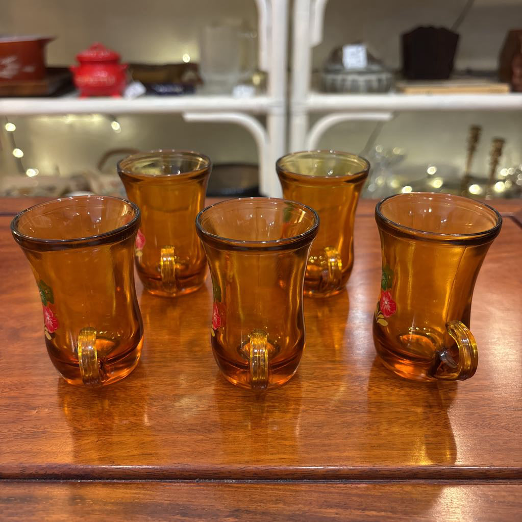 An amber glassware set with a decanter and 5 cordial glasses all with a floral pattern on the front sitting on a wooden table in a store setting. 