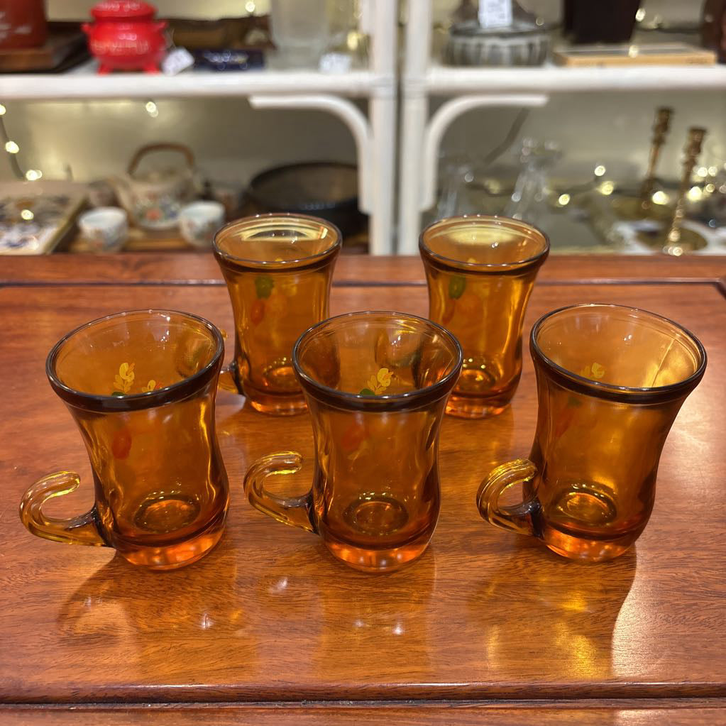 An amber glassware set with a decanter and 5 cordial glasses all with a floral pattern on the front sitting on a wooden table in a store setting. 