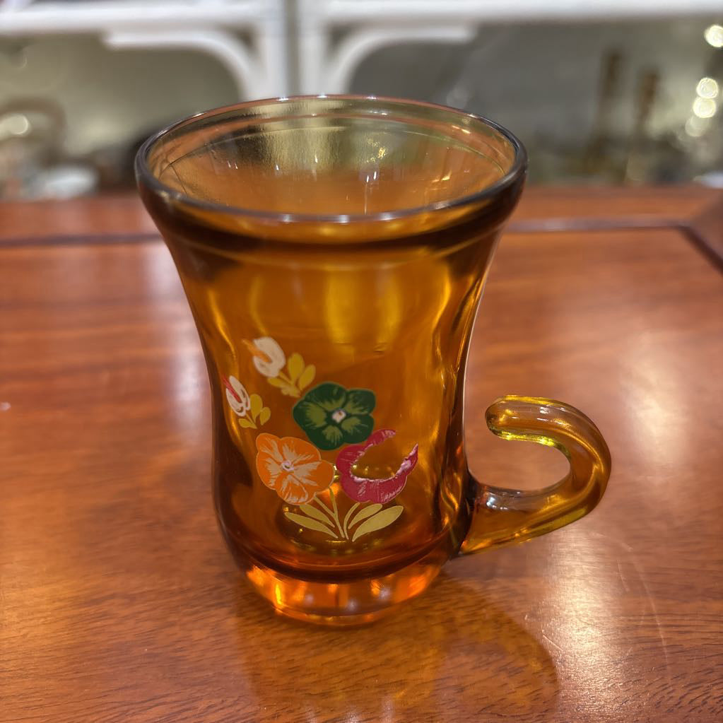 An amber glassware set with a decanter and 5 cordial glasses all with a floral pattern on the front sitting on a wooden table in a store setting. 