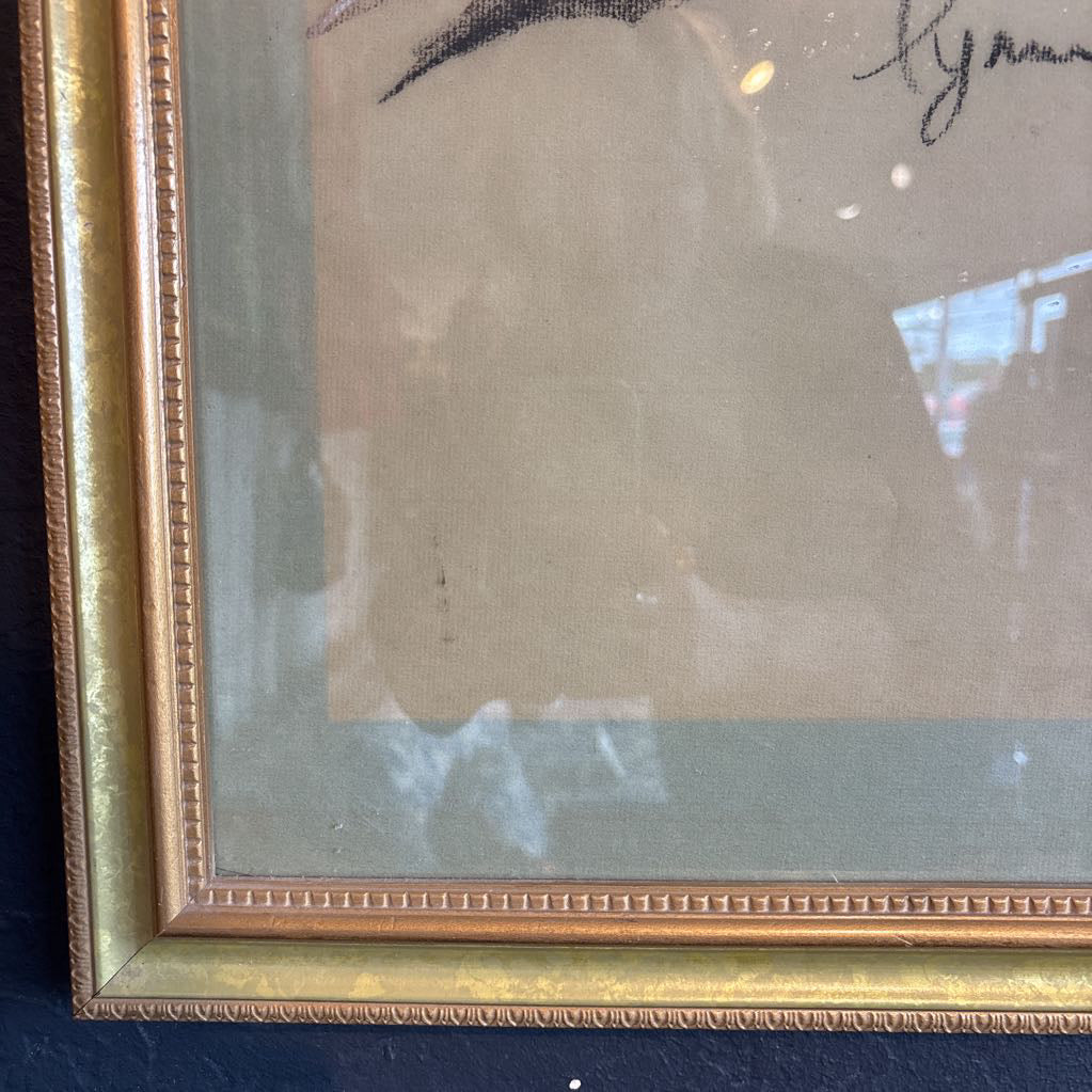 A vintage looking framed artwork of a blue and yellow Macaw on a branch with a signature under it. Its hanging on a black wall. 